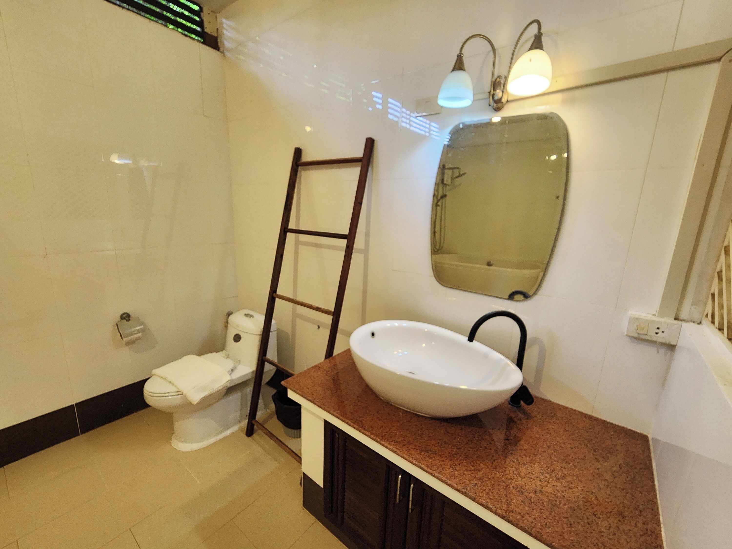Un baño con un inodoro blanco, un lavabo con encimera marrón, un espejo y una escalera apoyada en la pared.