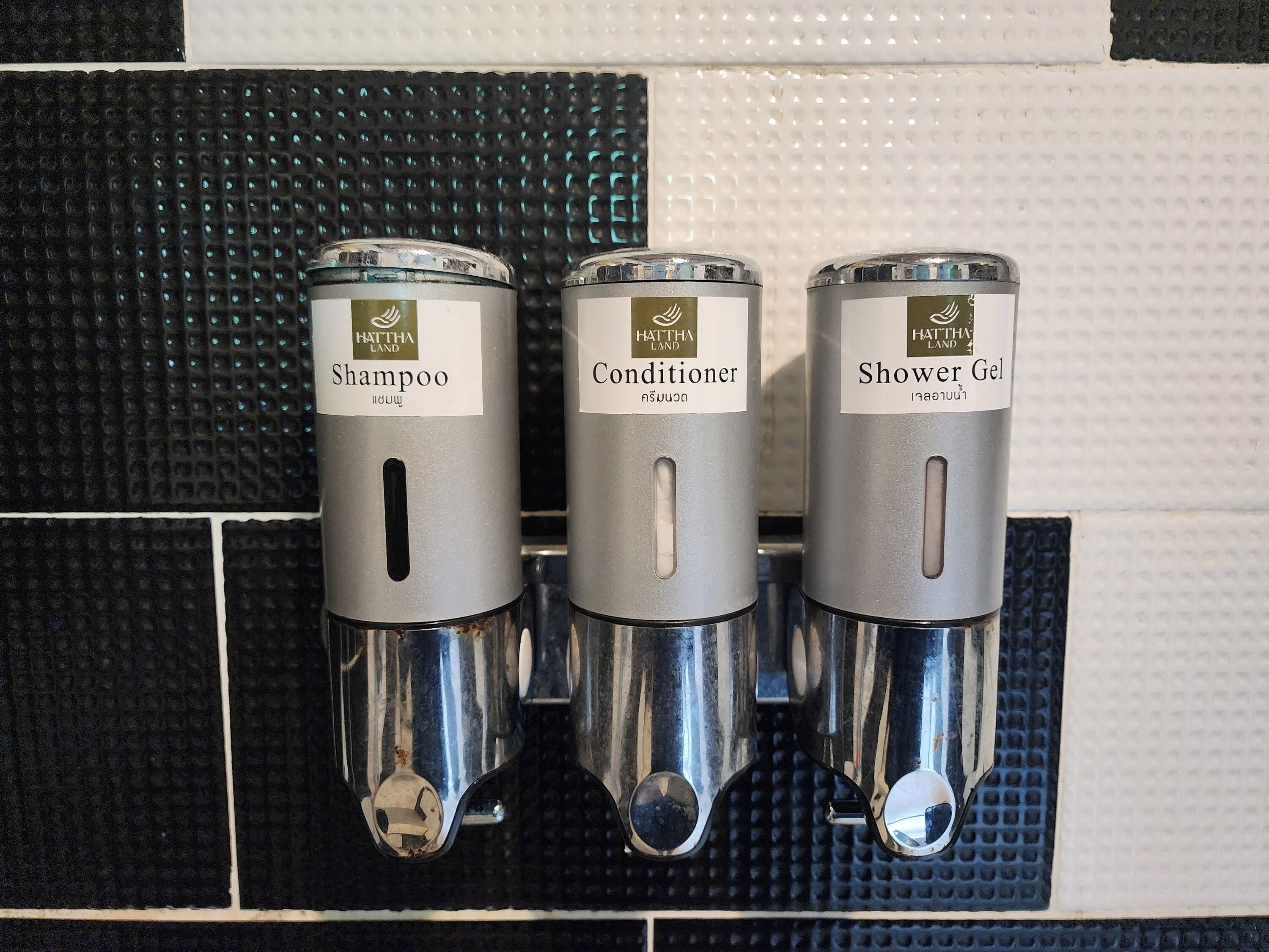 Three metal dispensers labeled 'Shampoo', 'Conditioner', and 'Shower Gel' mounted on a tiled wall.