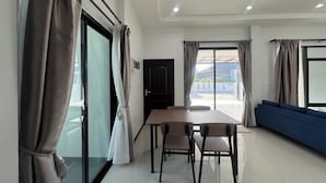 A modern living room with a dining area, large windows with curtains, and a blue sofa. - Poome House (Hat Yai)
