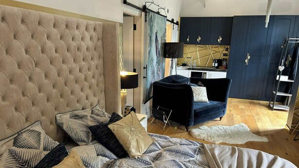 A bedroom with a tufted headboard, a bed with patterned bedding, a dark blue armchair, and a small kitchenette in the background. - Charming Shropshire Ground Level Barn (Bridgnorth)