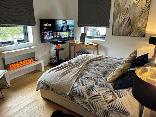 A modern bedroom with a large bed, a TV mounted on the wall, a fireplace, and a desk with a laptop.