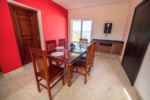 A dining area with a glass table, wooden chairs, a red wall, and a view through a window. - Queen's Hotel Mount Garden Kodaikanal (Kodaikanal)