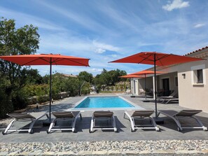 A swimming pool with lounge chairs and red umbrellas, surrounded by a garden and a building. - New Villa Floria**** with a private heated pool in southern Ardèche (Joyeuse)