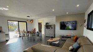 A modern living room with a sofa, dining table, and chairs. There is a kitchen area with cabinets and a TV mounted on the wall. - New Villa Floria**** with Private Heated Pool in South Ardèche (Joyeuse)