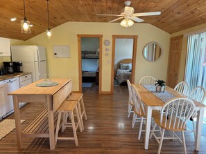 A cozy kitchen with wooden cabinets, a refrigerator, and a coffee maker. There is a dining area with a wooden table, benches, and white chairs. A ceiling fan is visible, and the room has wooden flooring.