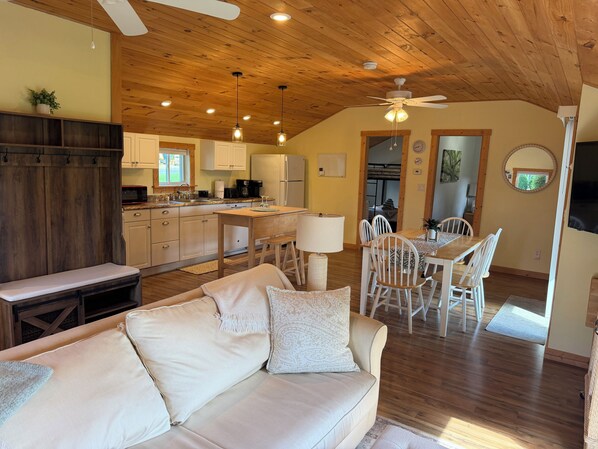 A cozy interior with a wooden ceiling, a dining area with white chairs, a kitchen with modern appliances, and a comfortable living space with a sofa.