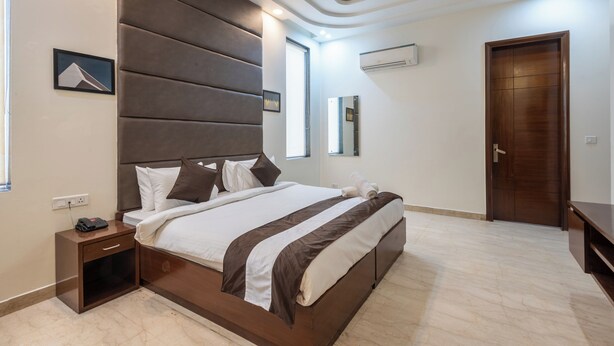 A modern bedroom with a large bed, a nightstand, a wooden door, and a wall-mounted air conditioner. - Saavi Hotel Sector- 46 (Gurugram)