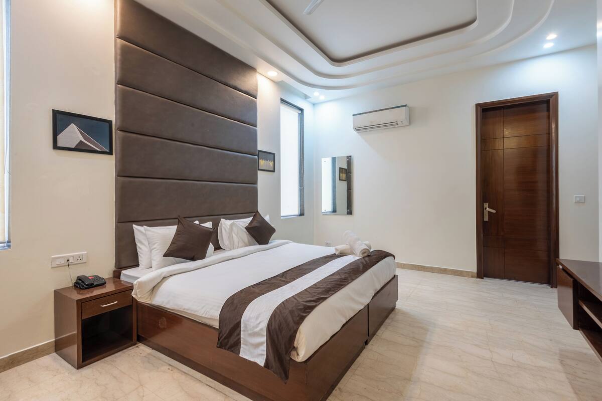 A modern bedroom with a large bed, a nightstand, a wooden door, and a wall-mounted air conditioner.