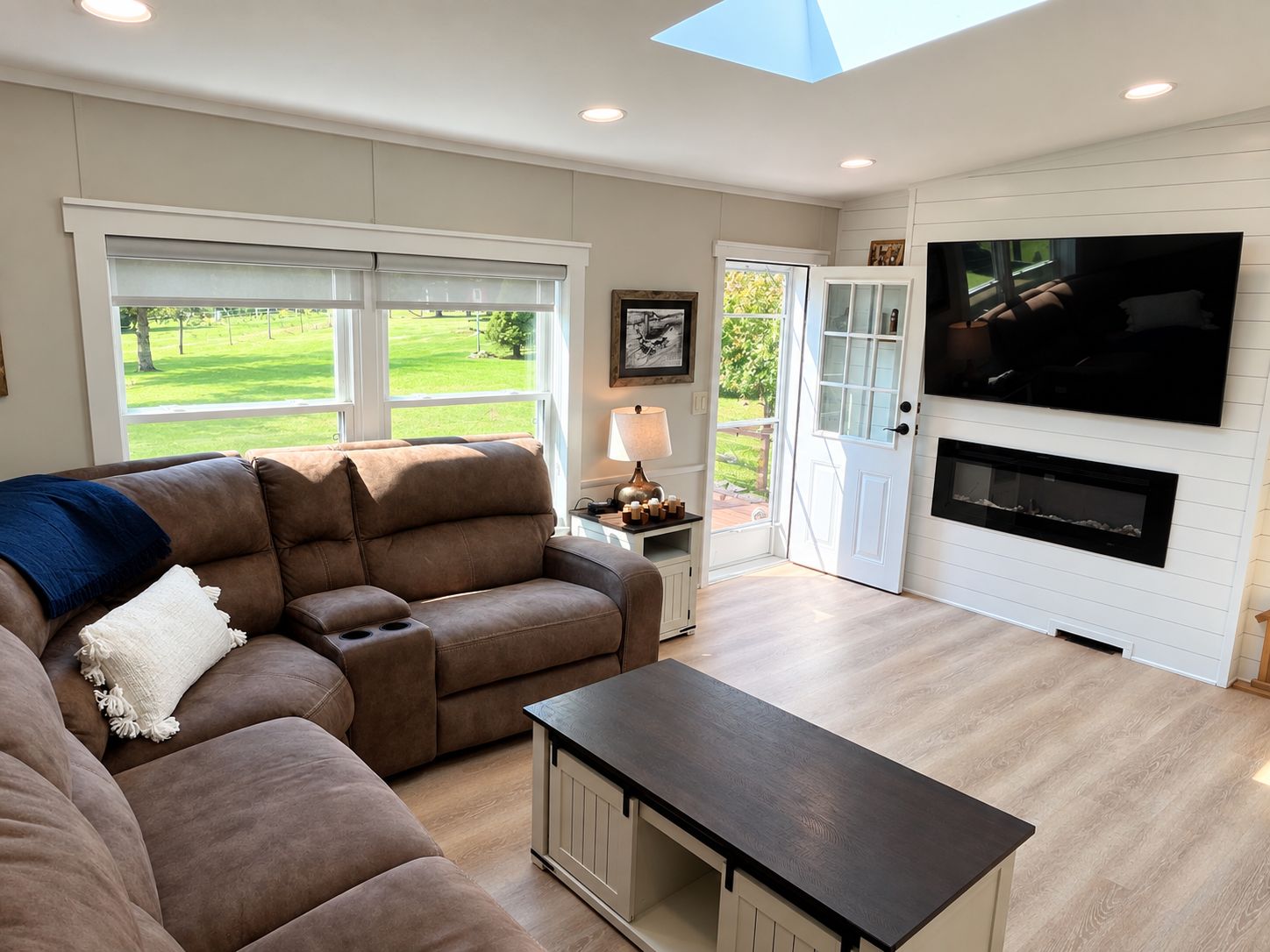 A living room with a sectional sofa, a coffee table, a fireplace, a TV mounted on the wall, and a large window with a view of a green lawn.