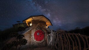 A wooden cabin with a circular red door, surrounded by a wooden fence, under a starry night sky. - Gaia" Hobbit House (Vittel)