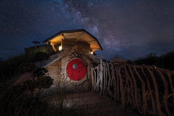 Une cabane en bois avec une porte rouge circulaire, entourée d’une clôture en bois, sous un ciel nocturne étoilé.
