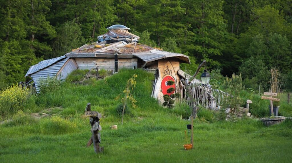 A rustic cabin with a sloped roof, surrounded by lush greenery and a small garden.