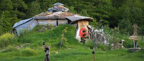 A rustic cabin with a sloped roof, surrounded by lush greenery and a small garden.