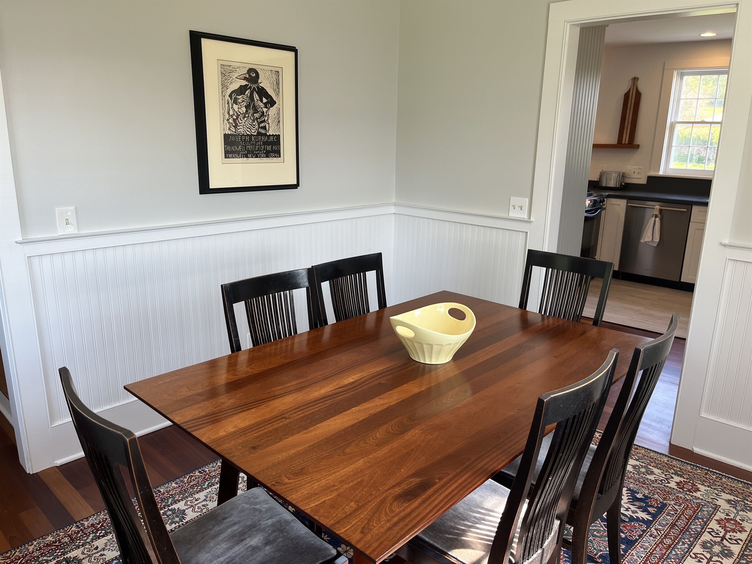 Una sala da pranzo con un tavolo e sedie in legno, un quadro appeso al muro e una cucina sullo sfondo.