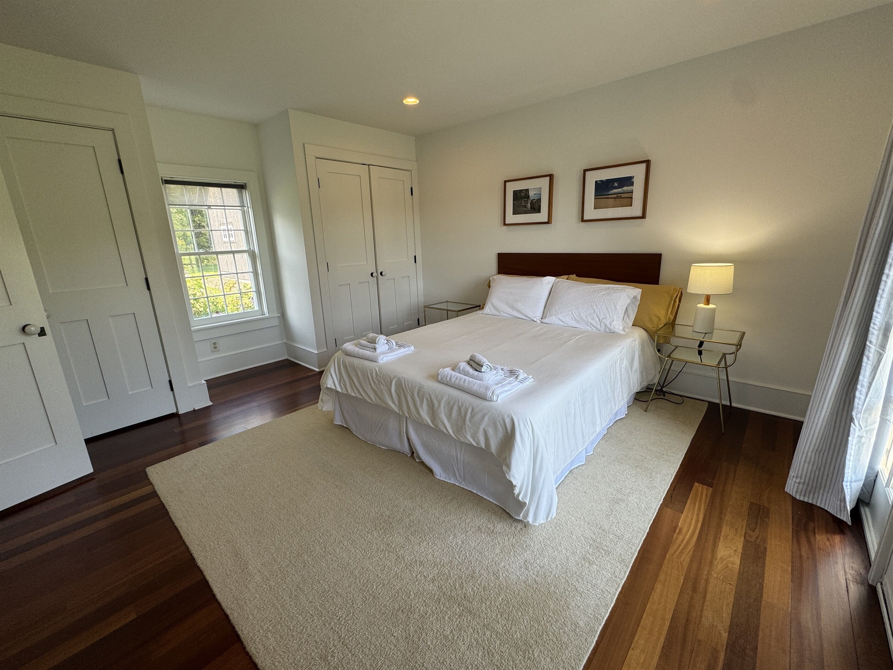 A neatly arranged bedroom with a large bed, a bedside table with a lamp, and two framed pictures on the wall.