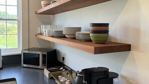 Cozinha com prateleiras de madeira que exibem tigelas e pratos, micro-ondas, cafeteira e janela com vista para o verde.
