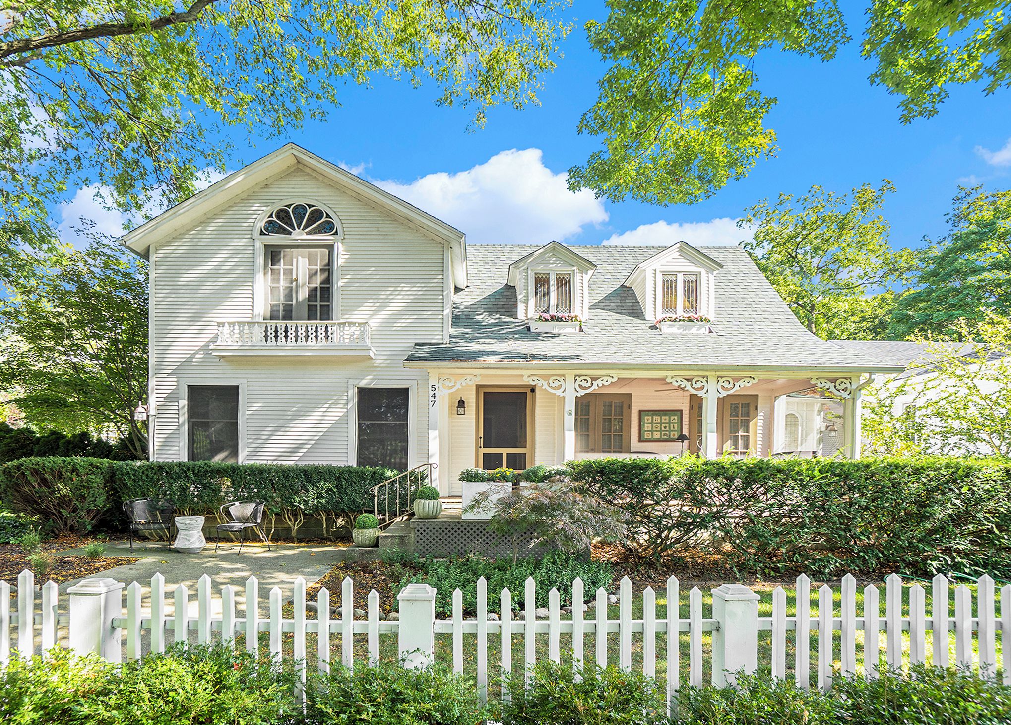 A white two-story house with a porch, a bay window, and a small garden area with potted plants and a bench.