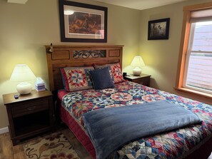 A bedroom with a wooden headboard, a quilted bedspread, two bedside lamps, a nightstand with a remote control, and a window with blinds.