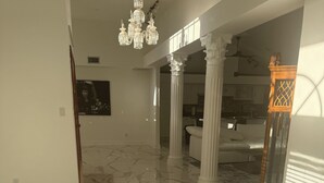 A spacious room with marble flooring, a chandelier, white columns, and a view into a kitchen area. - Mini Resort , 3 BR newly renovated, Gym , pool , spa , pickleball,basketball (Naples)