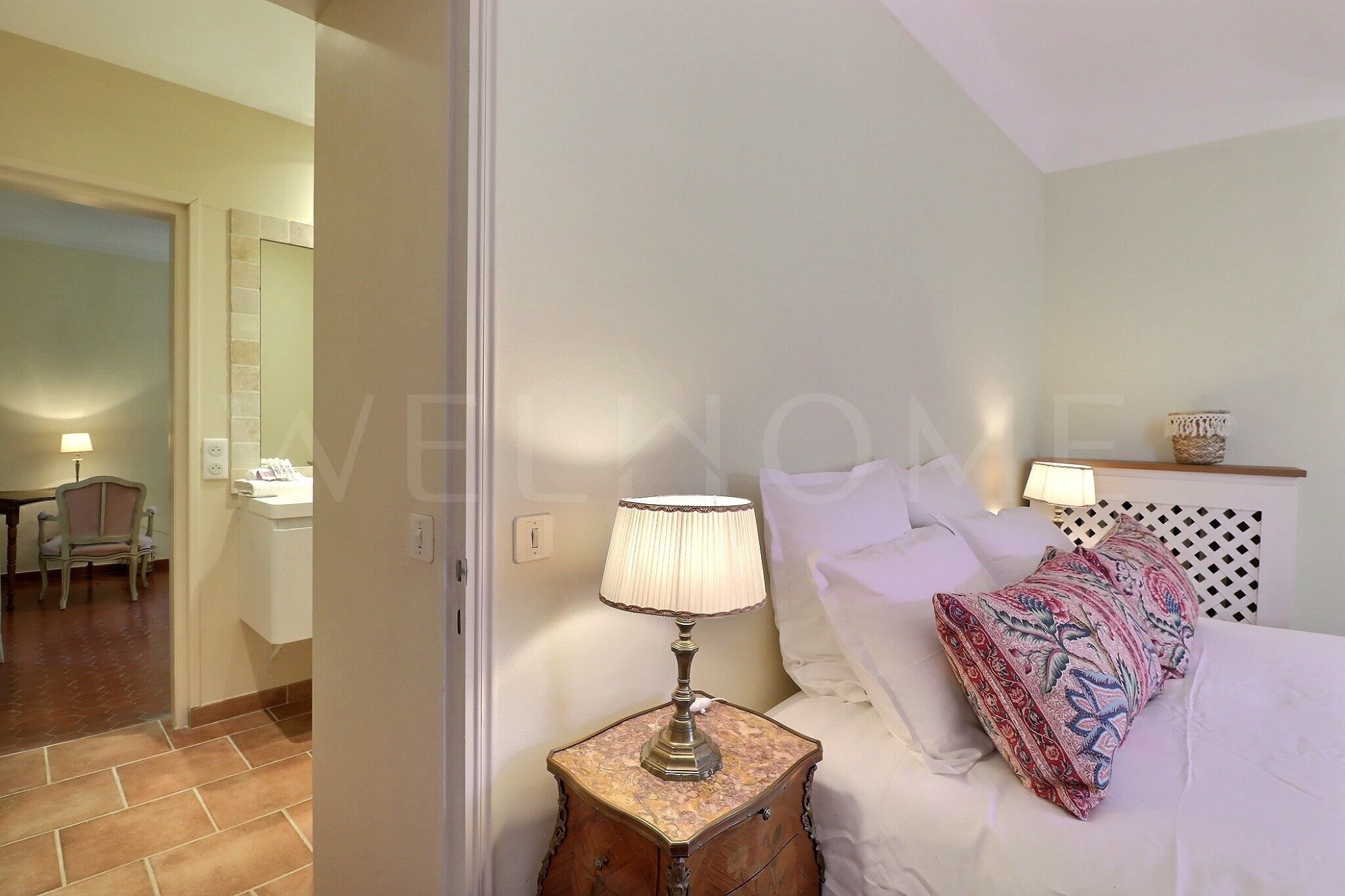 A bedroom with a bed, a nightstand with a lamp, a patterned pillow, and a chair in the adjacent room.