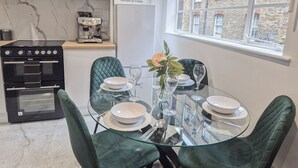 Une cuisine moderne équipée d’une cuisinière à gaz, d’un four et d’une machine à café. On y trouve une table à manger avec quatre chaises et un vase rempli de fleurs. Une fenêtre offre une vue sur un bâtiment en briques.