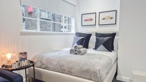 Une chambre à coucher bien rangée, avec un lit, des oreillers, une table de chevet avec une lampe et une fenêtre donnant sur un bâtiment en pierre.