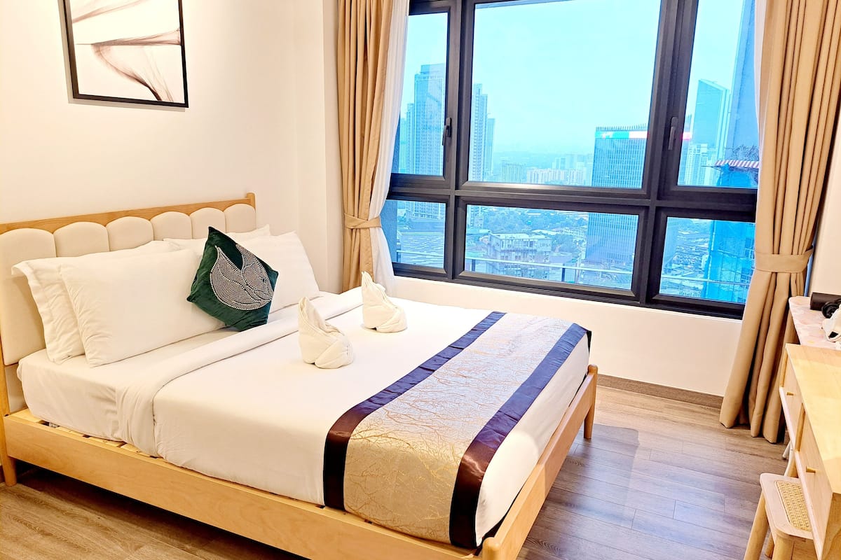 A neatly made bed with white and patterned linens, a green decorative pillow, and a pair of white slippers. A wooden headboard and a wooden desk with a chair are visible. The room has a large window with a view of a cityscape.