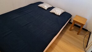 En säng med en mörkblå madrass och två kuddar, ett träbord som sängbord och en vit vägg.