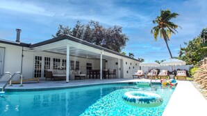 Una piscina con un porche cubierto, sillas de descanso y una palmera al fondo.