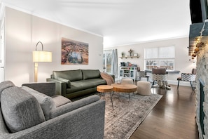 A living room with a green sofa, a lamp, a rug, and a dining area with chairs and a table. - Hillside Packers Haven: Hot tub, sleeps 14 (Green Bay)