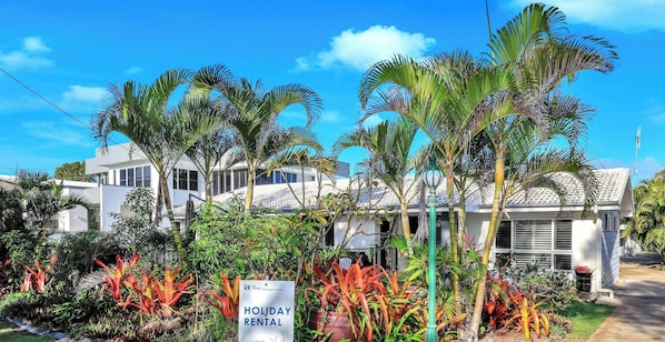 Un bâtiment moderne, sur lequel on peut lire une enseigne indiquant « Location de vacances », avec, au premier plan, une végétation tropicale luxuriante.
