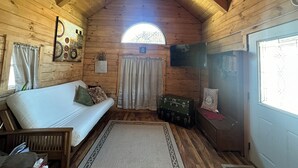 A cozy cabin interior with a wooden ceiling, a ceiling fan, a white sofa, a small rug, a window with curtains, and a vintage suitcase. - Cozy log cabin located in downtown Moravia, Iowa.  (Moravia)