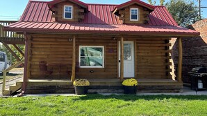 A wooden cabin with a red metal roof, two windows, and a front door. There are two potted plants on the porch. - Cozy log cabin located in downtown Moravia, Iowa.  (Moravia)
