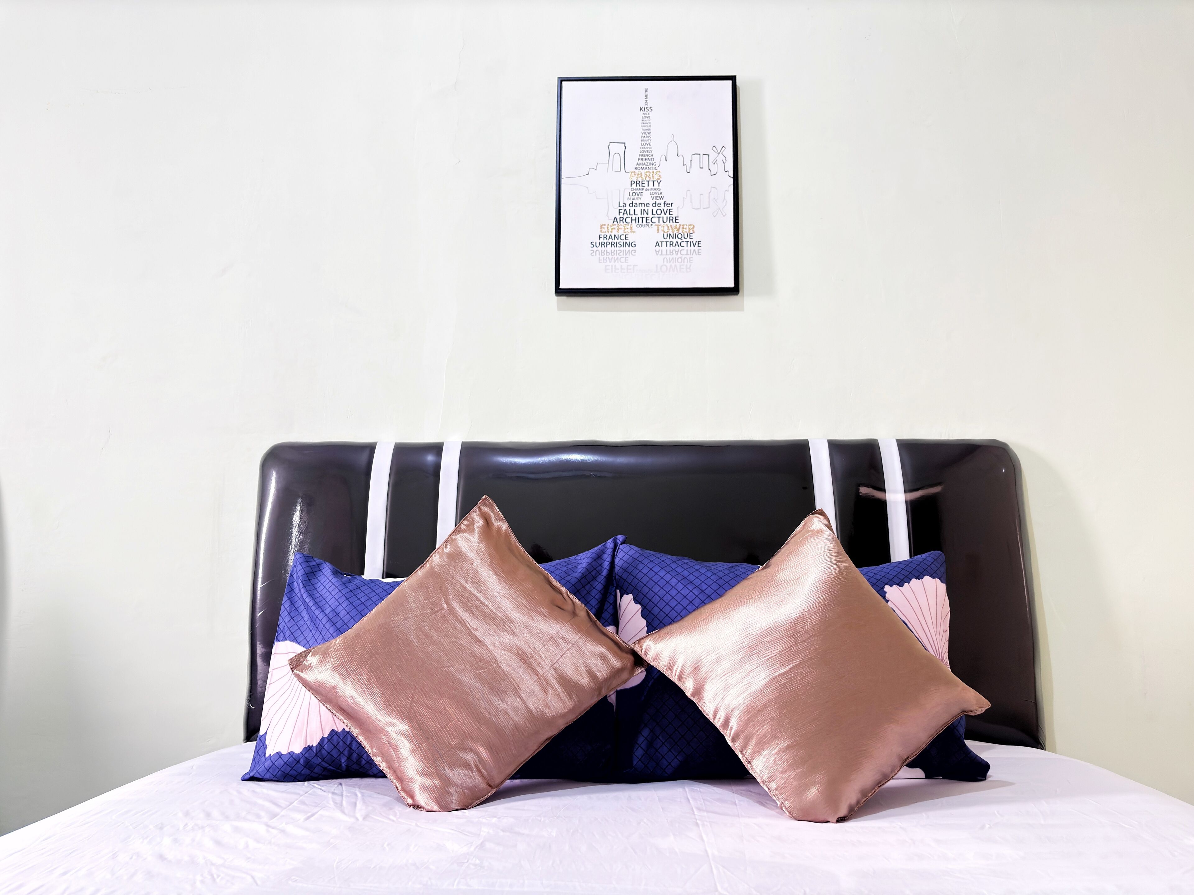 A bed with a black headboard, two rose gold pillows, and a framed picture of a city skyline on the wall.