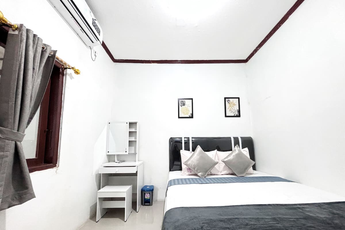 Quarto pequeno e limpo, com uma cama, uma escrivaninha e uma cadeira. Há uma janela com cortinas, um ar condicionado fixado na parede e duas fotos emolduradas.