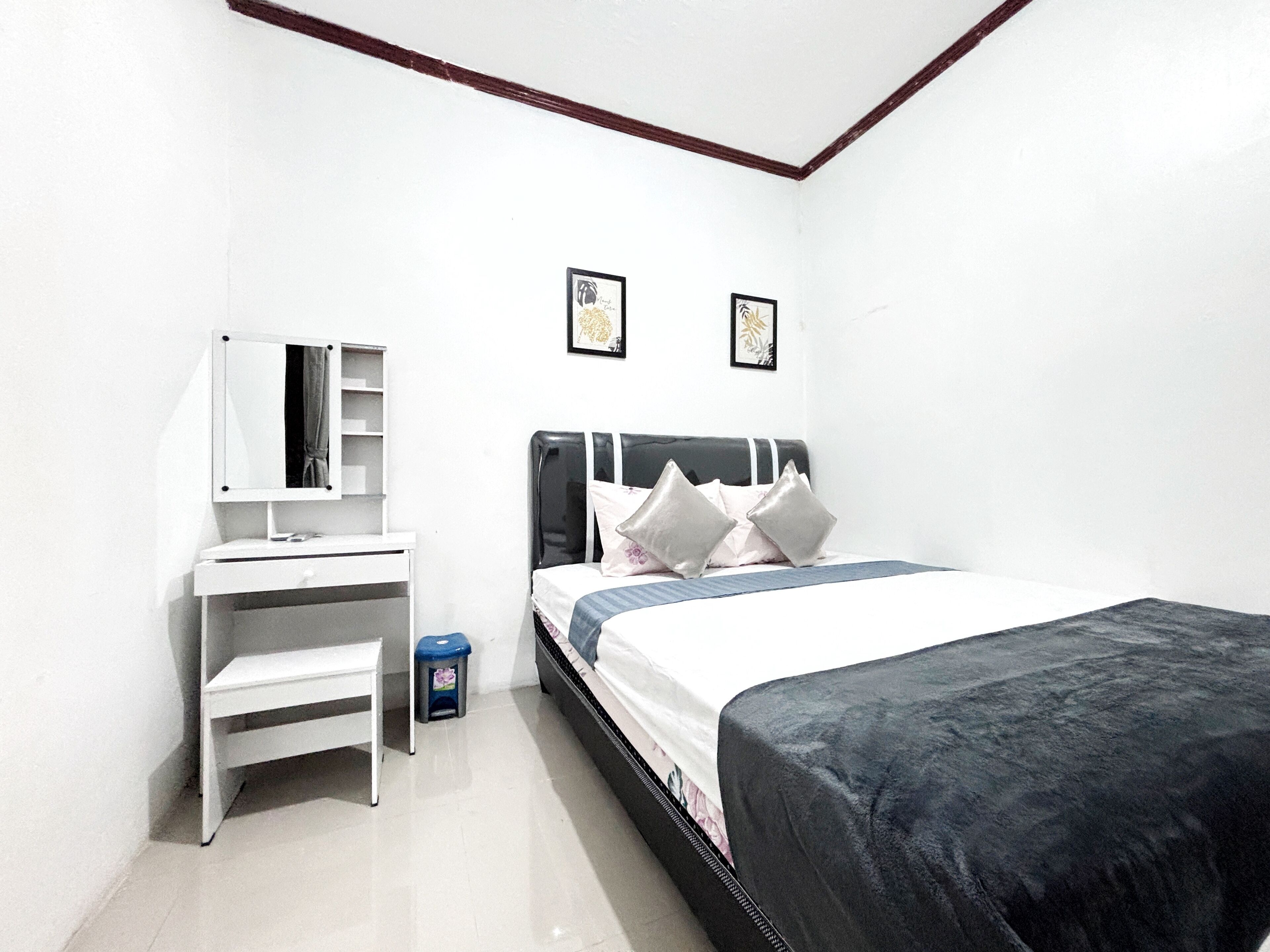 A bedroom with a bed, a desk, a chair, a trash bin, and two framed pictures on the wall.