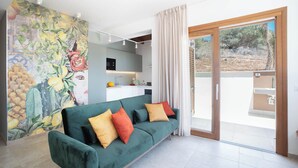 A modern living room with a green sofa, colorful cushions, a kitchen in the background, and a sliding glass door leading to an outdoor area. - Villa Eos  (Castellammare del Golfo)