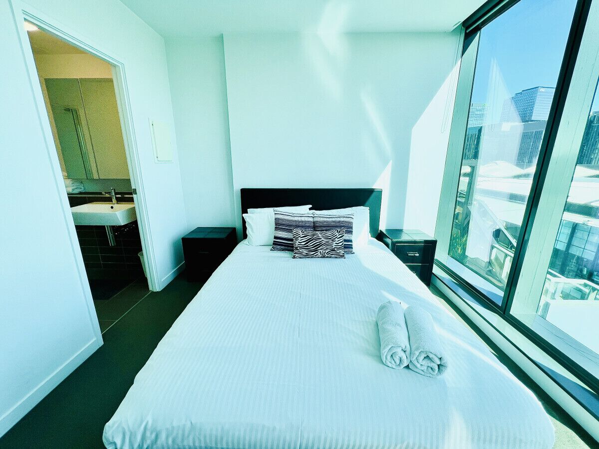 Hotelkamer met een groot bed, een nachtkastje, een raam met uitzicht op de stad en een zichtbare badkamer door een open deur.