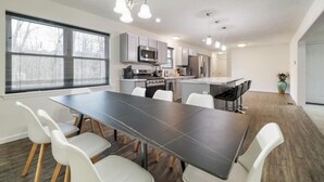 A modern kitchen with stainless steel appliances, a dining area with a dark table and white chairs, and a large window with blinds. - Pocono Mountain Escape! Fire pit•Hot tub•Games•Pool•Lake•Fishing•Golf•Ski•Hiking (Tobyhanna)