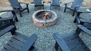 A circular outdoor seating area with four Adirondack chairs around a stone fire pit. - Pocono Mountain Escape! Fire pit•Hot tub•Games•Pool•Lake•Fishing•Golf•Ski•Hiking (Tobyhanna)
