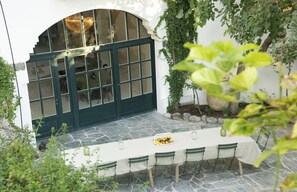 Un espace de restauration en plein air doté d’un sol en pierre, d’une longue table dressée pour six personnes et offrant une vue sur une pièce avec une grande fenêtre et un lustre.