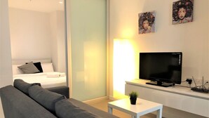 Quarto de hotel moderno com sofá cinza, cama com roupa de cama branca, televisão de tela plana e pequena mesa com uma planta.