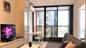 Uma sala de estar moderna com televisão de tela plana, um sofá, uma mesa pequena e vista para a cidade através de grandes janelas.