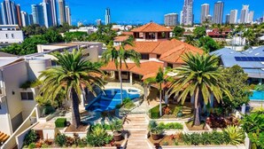 A residential area with a swimming pool, palm trees, and a city skyline in the background. - Main River Grandeur Mansion, 5 Bedroom, Pool & Sauna (Surfers Paradise)