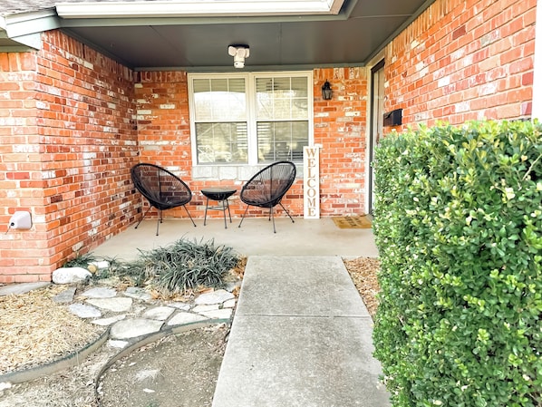 A brick exterior with a welcome sign, two chairs, and a hedge.