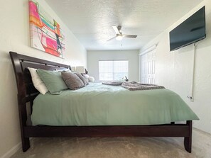 A bedroom with a bed, a ceiling fan, a TV mounted on the wall, and a window with blinds.