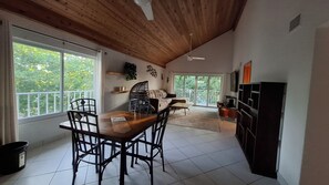 A dining area with a wooden table and chairs, a sofa, and a view through a large window. - 2 Bed Penthouse on Seven Mile Beach. Private Beach Cove, & 3 Pools! (#7) (Grand Cayman)