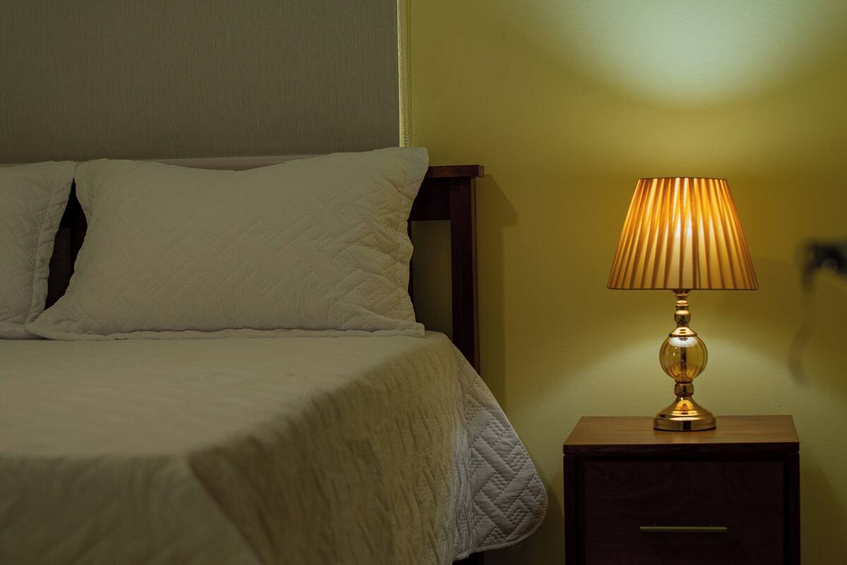A neatly made bed with a white quilt, a bedside table with a lamp, and a wooden nightstand.