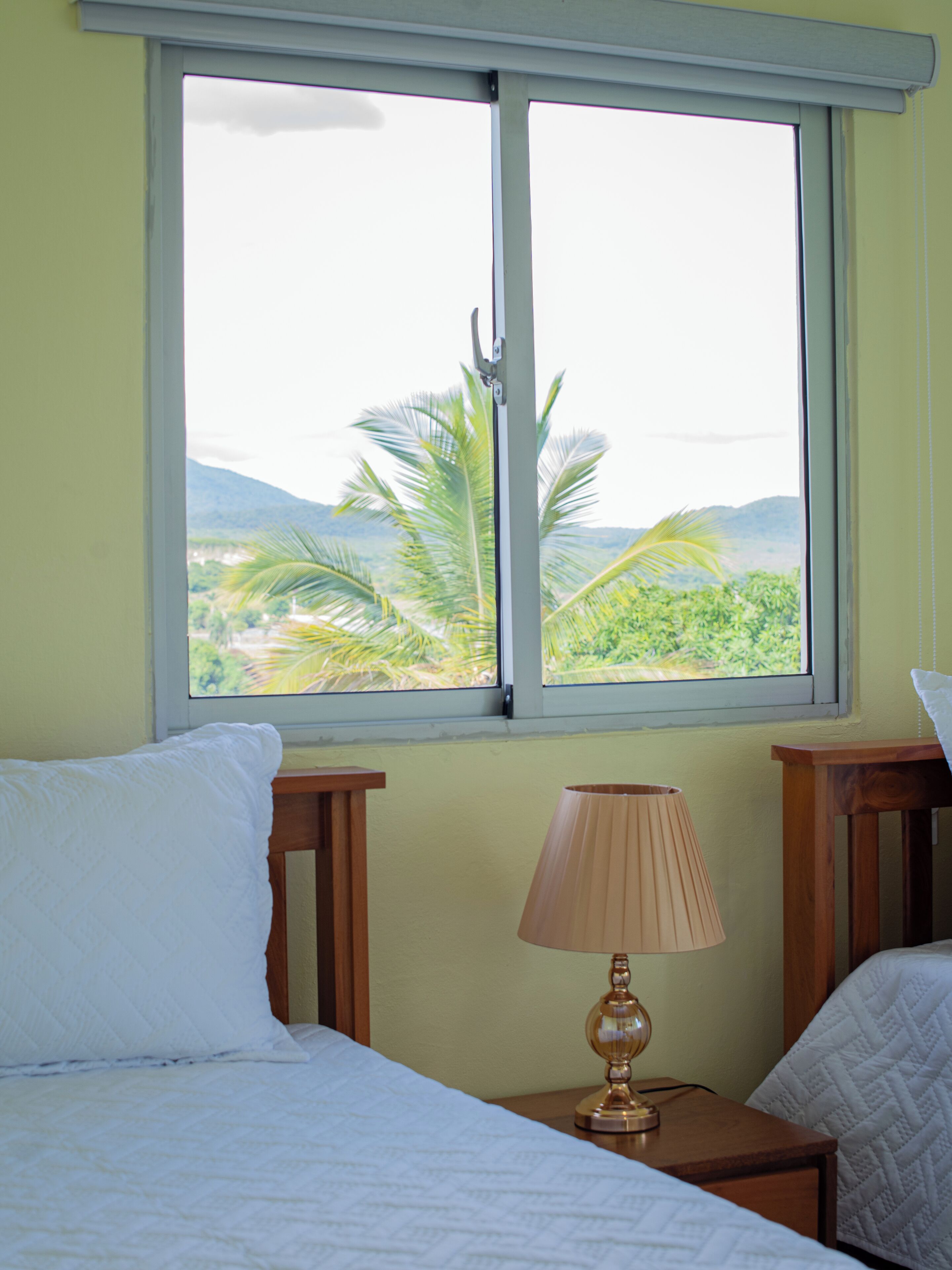Ein Zimmer mit einem Bett, einem Nachttisch mit Lampe und einem Fenster mit Blick auf Palmen und Berge.
