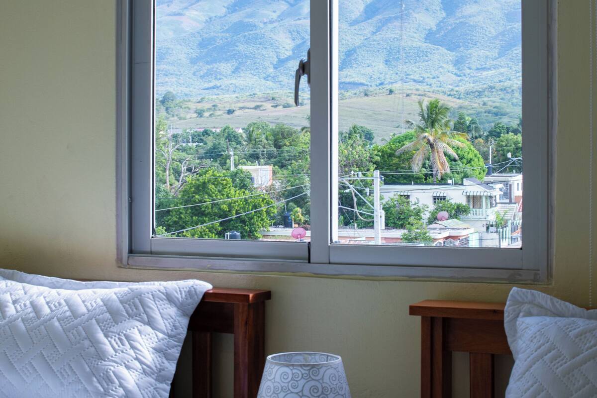 A room with two beds, a window with a view of mountains and a town, and a bedside table with a lamp.